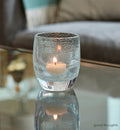 good thoughts, transparent light blue with small bubble pattern hand-blown glass votive candle holder on a reflective coffee table with gray couch in background.