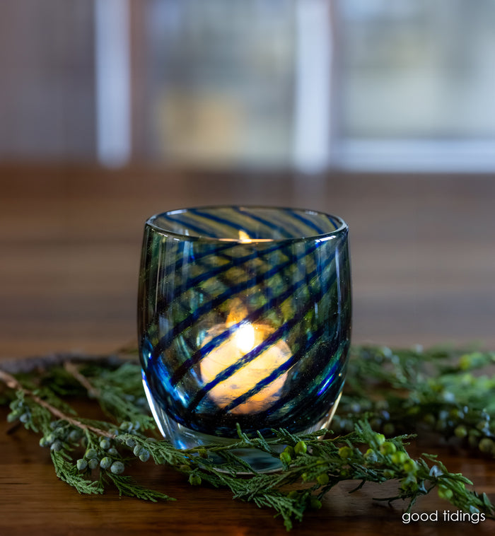 blue swirls wrapped around clear glassybaby hand-blown glass candle holder with lit tealight. green garland around it on wood table.