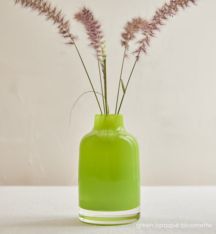 green opaque bloomette, lime green opaque hand-blown glass flower bud vase with purple grass  in front of a cream background.
