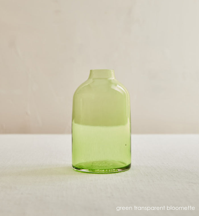green transparent bloomette, transparent green hand-blown glass flower bud vase on a cream table in front of cream wall.