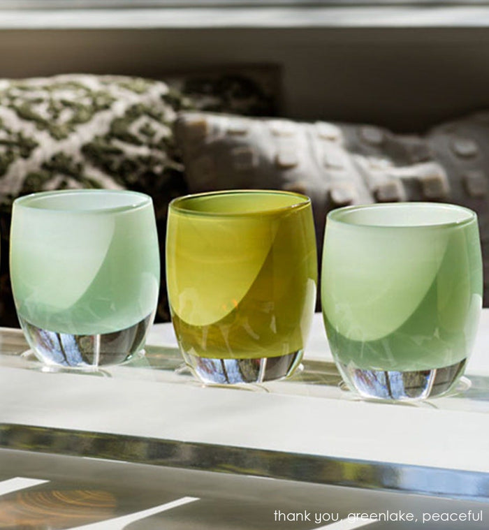 greenlake, olive green hand-blown glass votive candle holder with peaceful and thank you on a glassybaby baby acrylic baby stand sitting in the sunshine.