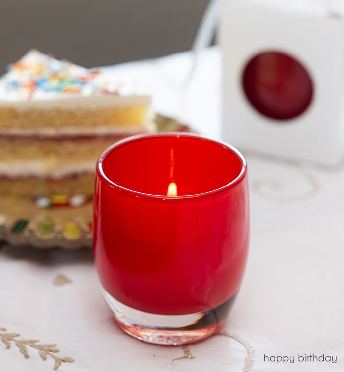 happy birthday bright red, hand-blown glass votive candle holder with a blurred birthday cake in the background