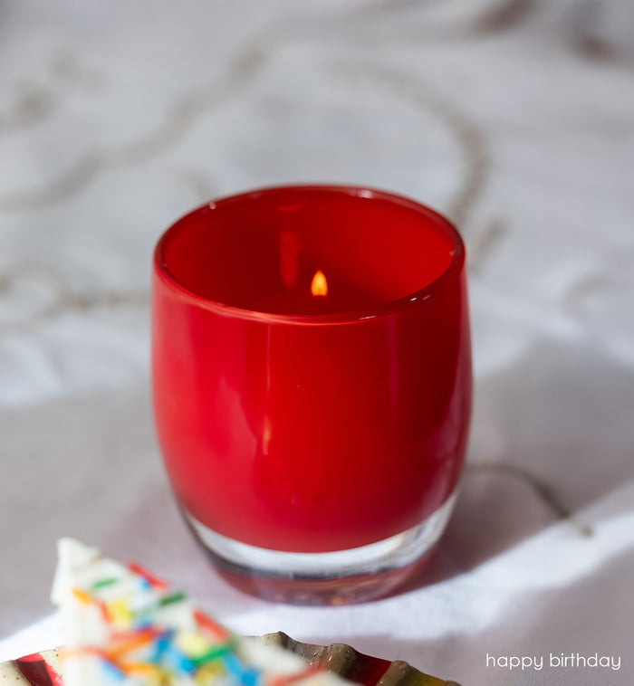 happy birthday bright red, hand-blown glass votive candle holder with a blurred birthday cake in the background