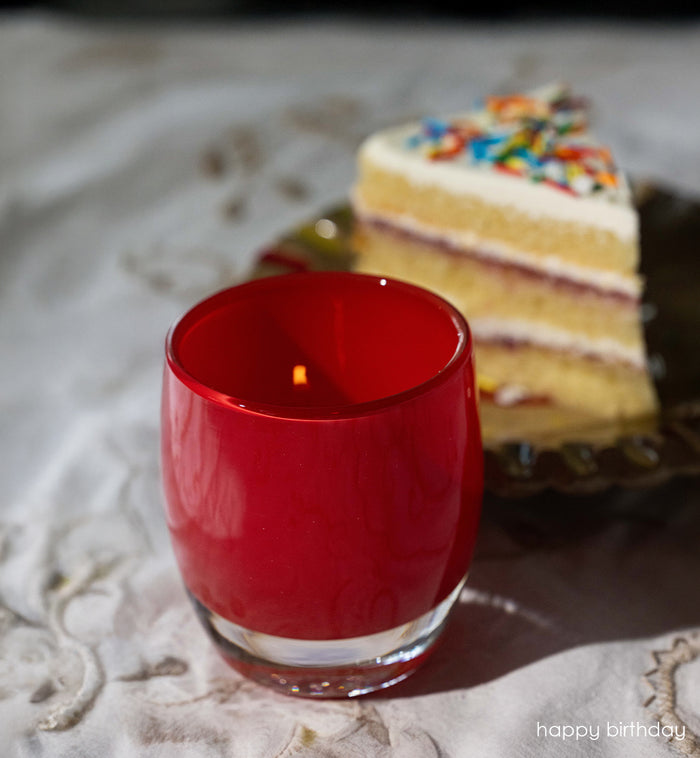 happy birthday bright red, hand-blown glass votive candle holder with a blurred birthday cake in the background