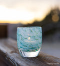 This image features a beautifully crafted glass candle holder, with a unique design that resembles fluffy white clouds against a light blue background. The holder is filled with a tealight candle, providing a soft, warm glow that enhances the glass's intricate details.