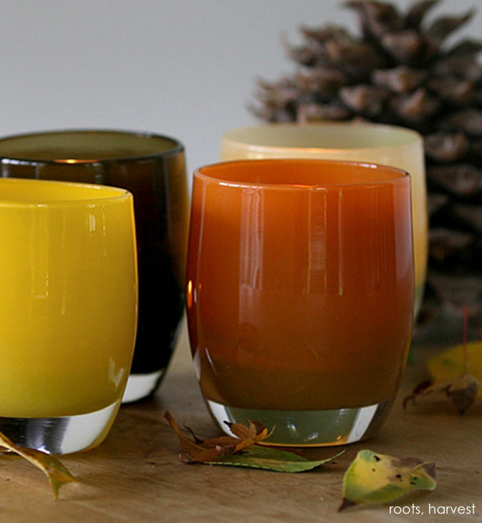 harvest deep orange hand-blown glass votive candle holder. Paired with roots on a table with fall leaves and a pinecone.