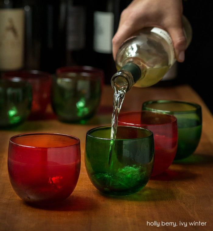 several holly berry drinkers transparent green hand-blown drinking glasses with ivy winter drinkers on a wood table with wine being poured into each.