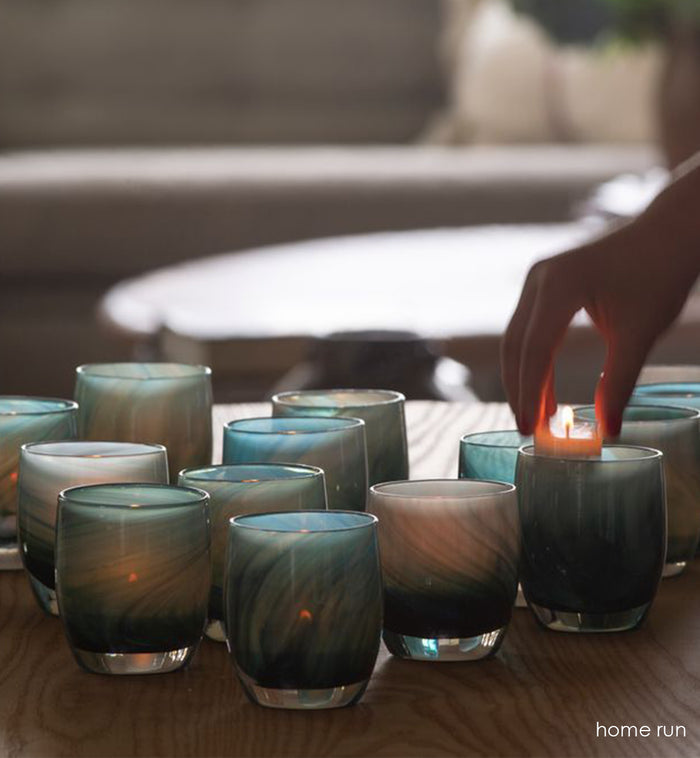 grouping of home run, multi-colored blue swirl hand-blown glass votive candle holders on a light wood coffee table, with a tealight being placed inside one.