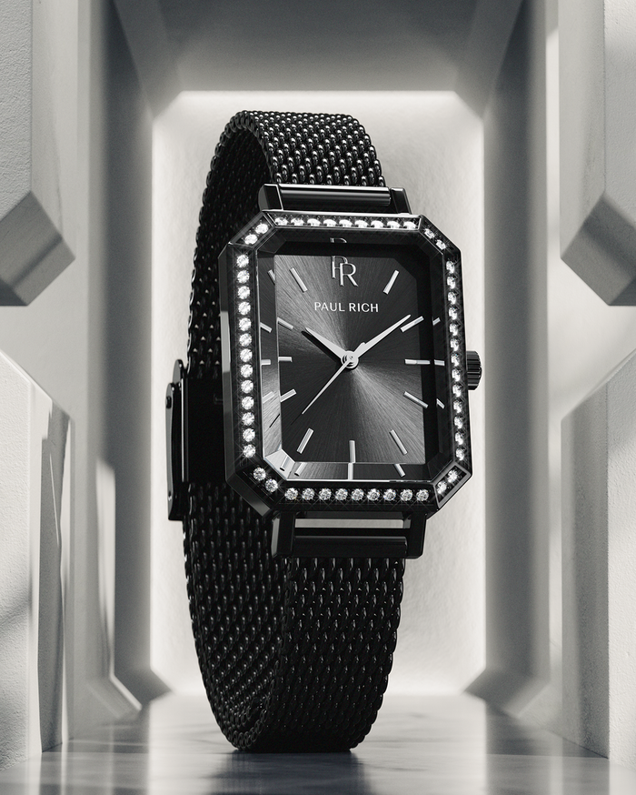 Black Paul Rich watch with black sunray dial, gem-set octagonal bezel, and black mesh strap, displayed upright in a geometric white architectural setting with dramatic lighting.
