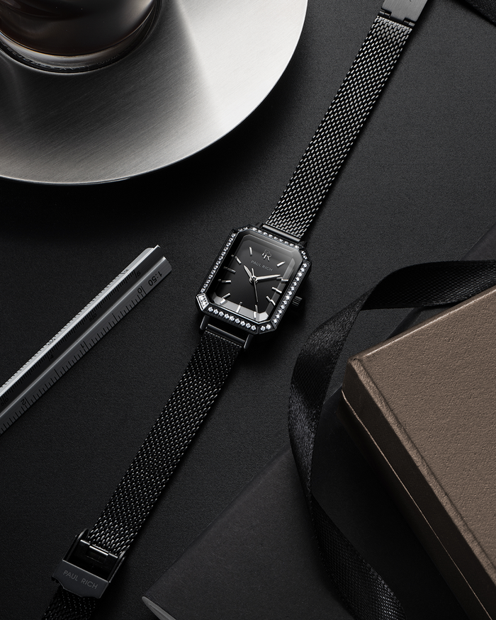 Black Paul Rich watch with black sunray dial, gem-set octagonal bezel, and black mesh strap, displayed flat on a dark surface alongside a cup of coffee, a ruler, a notebook, and a black ribbon.