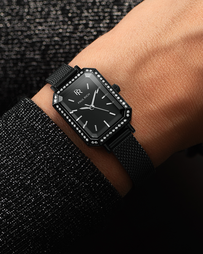 Black Paul Rich watch with black sunray dial, gem-set octagonal bezel, and black mesh strap, worn on wrist with a sparkling black sleeve.
