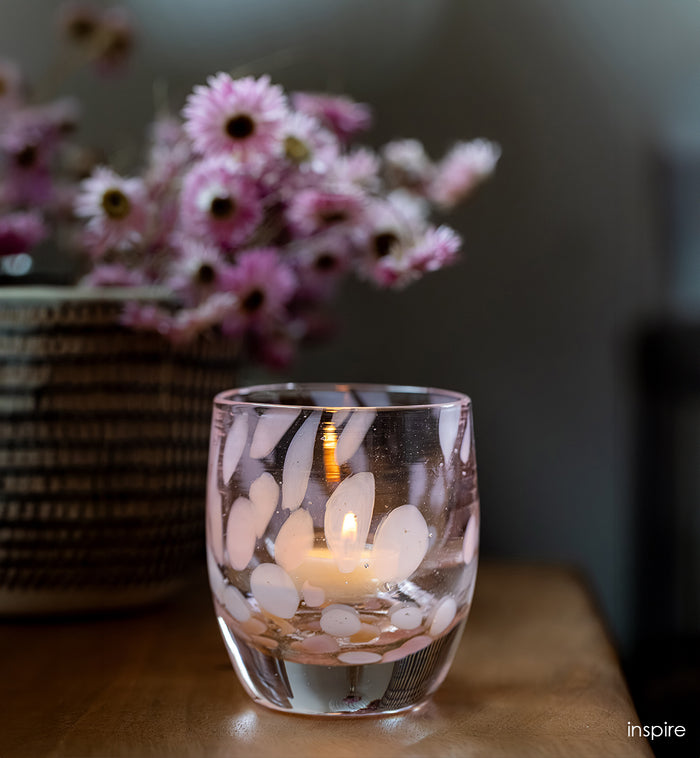 pink transparent glass candle holder with a lit candle on a wooden surface, with pink flowers in the background.