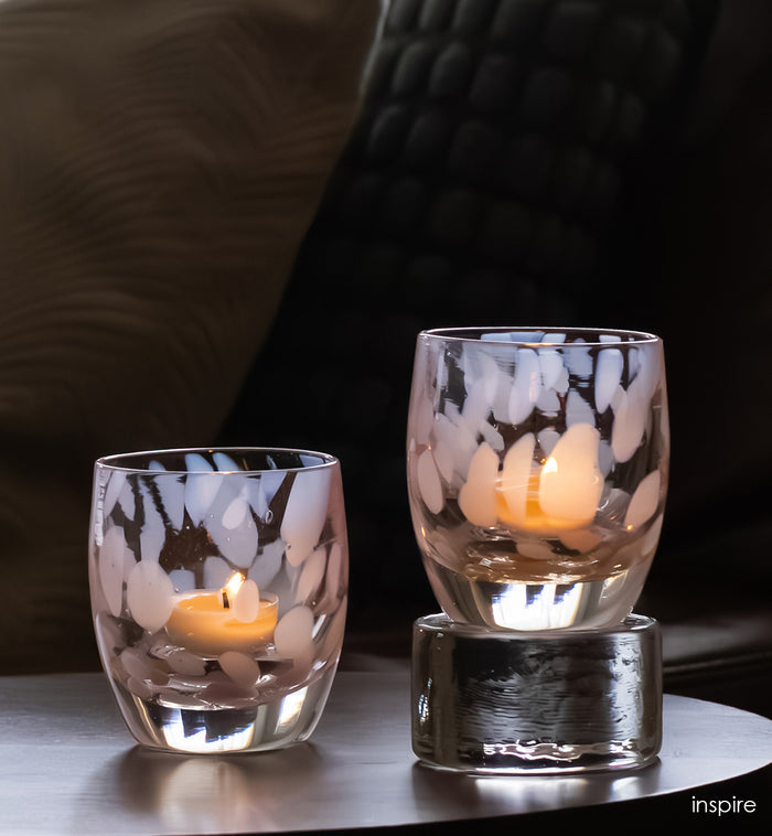 Two pink and clear glass candle holders with candles on a reflective surface, blurred background
