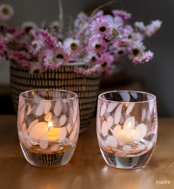 Two glass candle holders with lit candles on a wooden surface, with a basket of pink flowers in the background.