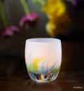 intuition blue, green and pink petals of color at the base of a white, hand-blown glass votive candle holder on a wooden surface, blurred flowers in the background