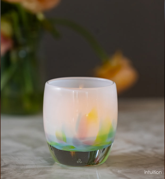 intuition blue, green and pink petals of color at the base of a white, hand-blown glass votive candle holder on a surface, blurred flowers in the background