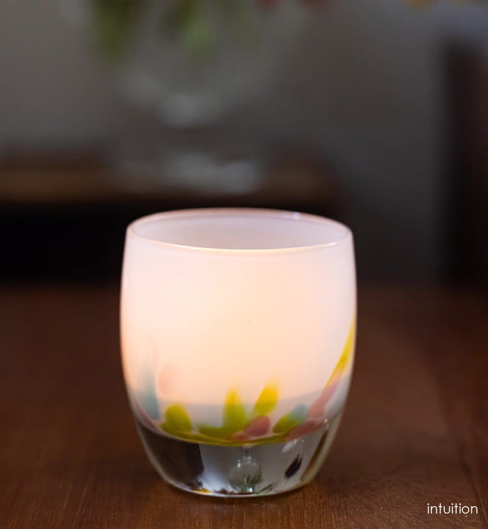 intuition blue, green and pink petals of color at the base of a white, hand-blown glass votive candle holder on a wooden surface