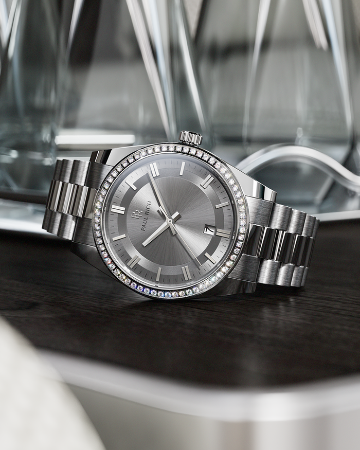 Silver-tone Paul Rich watch with silver sunray dial, diamond-set bezel, and silver link bracelet, displayed on a dark wooden surface with reflective glass decanters in the background.