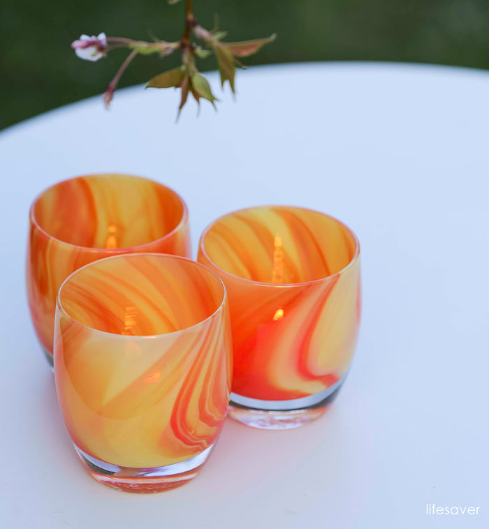 three lifesaver orange and yellow swirled, hand-blown glass votive candle holders on an outdoor table with a white tablecloth..