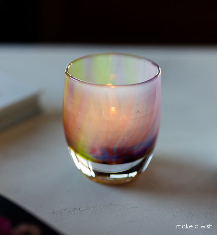 Multicolored glass candle holder with a lit candle on a white surface.