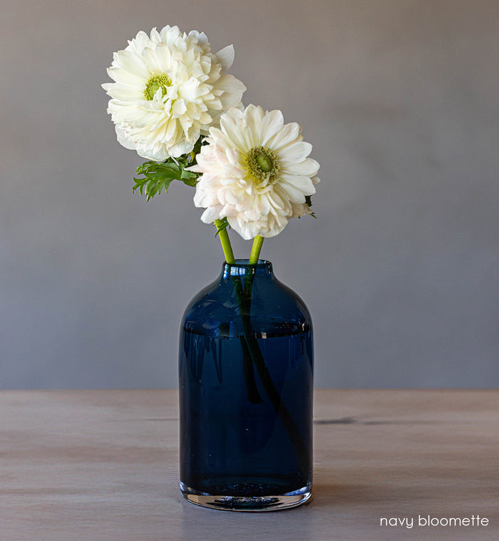 transparent navy hand-blown glass flower bud vase with white flowers on wood table and gray background