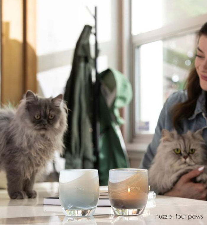 etched four paws, dark foggy gray hand-blown glass votive candle holder with 'Penny' on it. Paired with Cleo on a white table with two kitties, Penny and Cleo in the background.