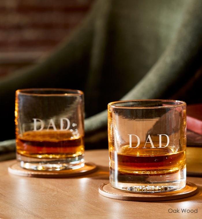 two Oak Wood rockers, light brown hand-blown glass lowball drinking glasses. with the word dad custom etched on two leather coasters atop a wood table with green chair in background.