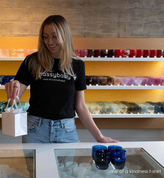 annie wearing a glassybaby one of a kindness t-shirt while holding a wrapped glassybaby in a store.