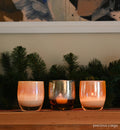 precious cargo is a beautiful set of 3 hand-blown glass votive candle holders; frankincense, gold, and myrrh. sitting atop a light wood mantle with framed wall art and decorative evergreen branches behind.