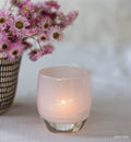 soft pink candle in a glass holder with pink flowers in the background on a light surface.
