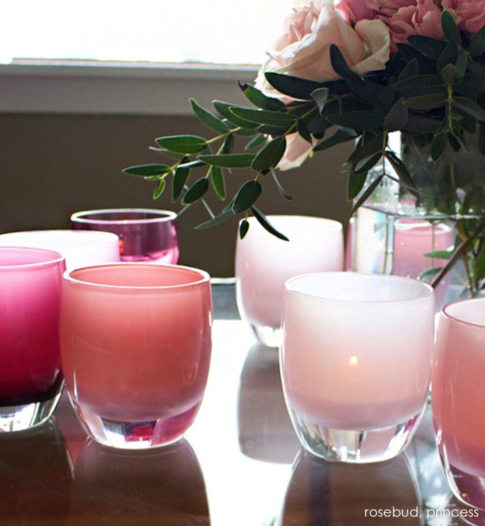 princess soft pink hand-blown glass votive candle holder grouped with rosebud on a glass table with a vase of flowers.