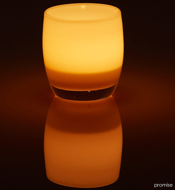promise cream hand-blown glass votive candle holder lit on a dark reflective surface.