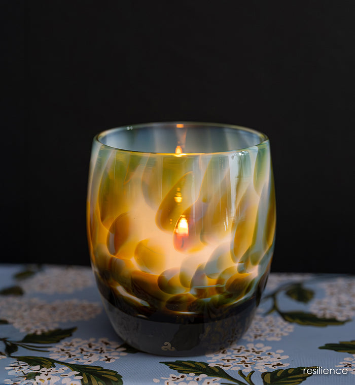 resilience | white hand-blown glass candle holder with brown, blue, and green petal deign with a lit candle against a dark background
