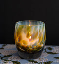 resilience | white hand-blown glass candle holder with brown, blue, and green petal deign with a lit candle against a dark background