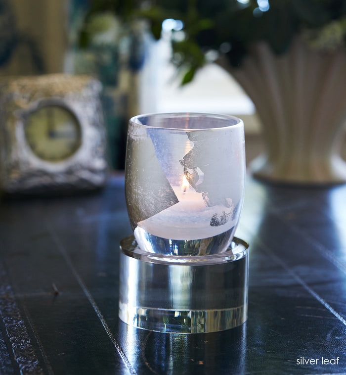 silver leaf, hand-blown glass votive candle holder with silver leaf, with a lit tealight glowing inside. sitting on a round baby stand on a navy blue desk with decorative clock and vase in background.