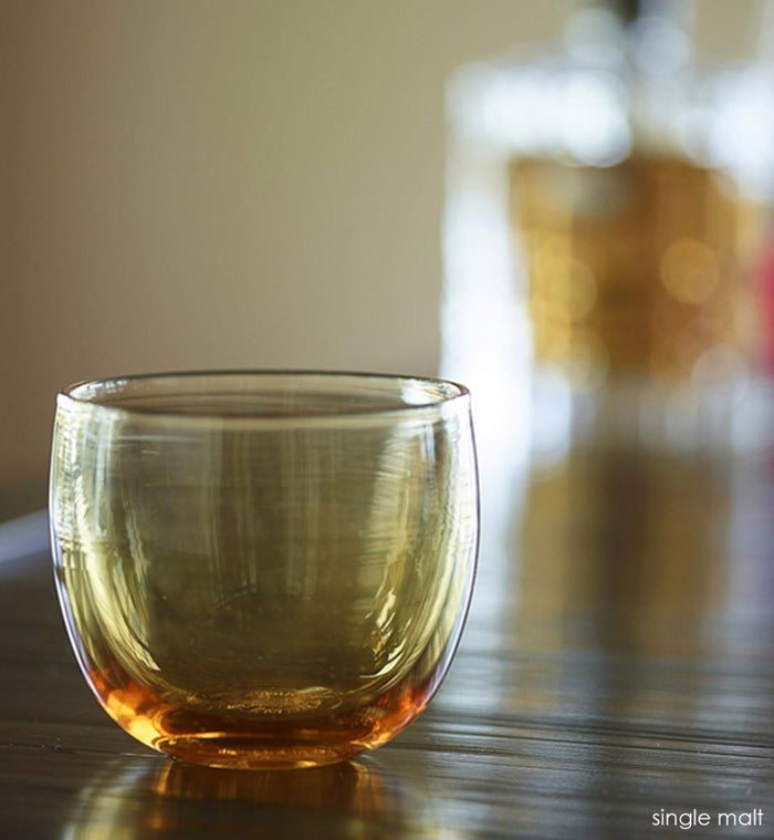 single malt drinker, transparent light amber hand-blown drinking glass on a dark wood surface with a liquor bottle blurred in background.
