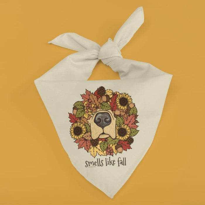A white fabric dog bandana with a tied top, featuring a printed graphic of a dog face surrounded by autumn leaves, sunflowers, and acorns, with the text 'smells like fall' below, set against a solid mustard yellow background.