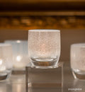 several snowglobe hand-blown white frosted glass votive candle holders, on square baby stand and gray surface with gold frame blurred in background.