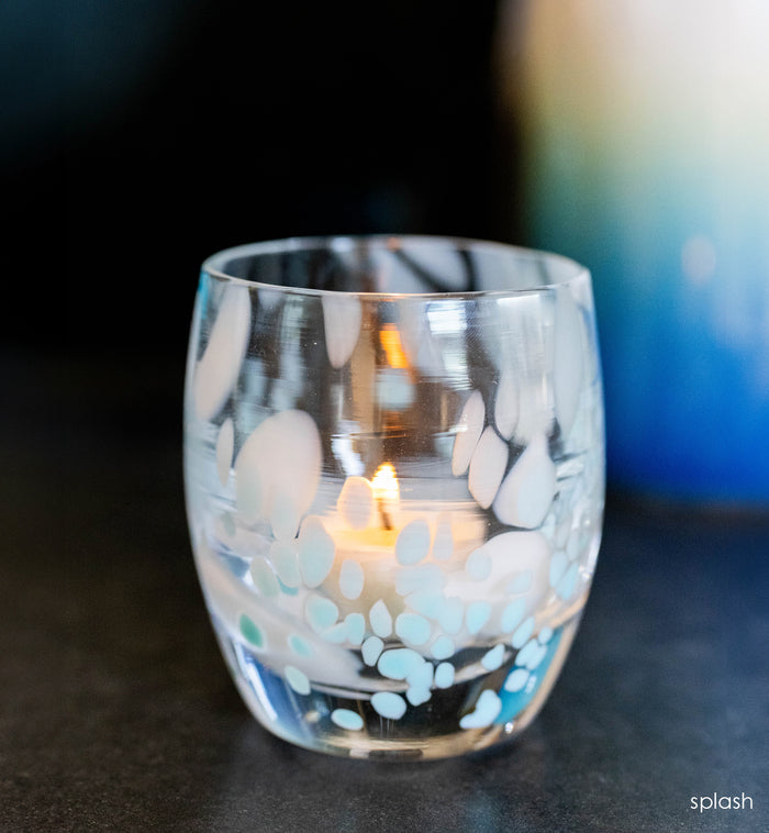 Clear glass candle holder with white and blue spotted design containing a lit candle on a dark surface.