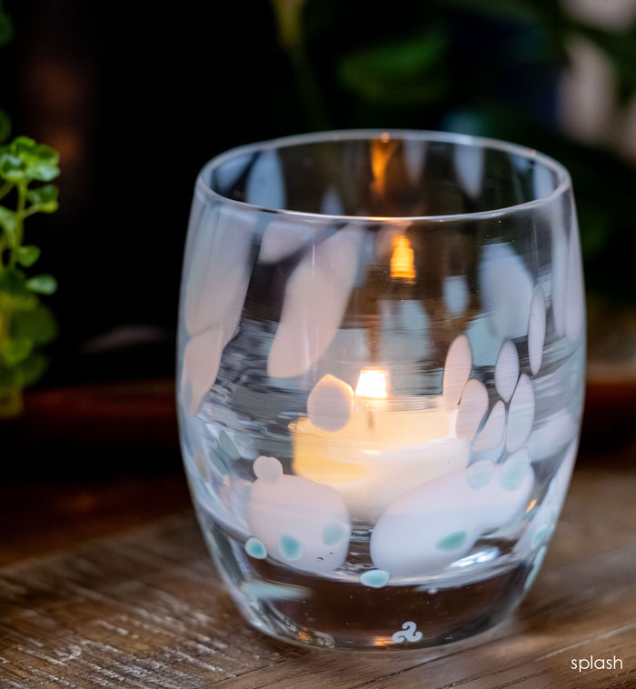 Clear glass candle holder with white and blue spotted design containing a lit candle on a wooden surface, blurred greenery in the background.