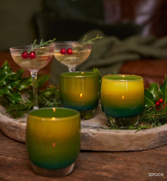 three spruce, forest green opaque hand-blown glass votive candle holders on a wood table and tray with lit tealights inside, next to holly plants and festive cocktails.