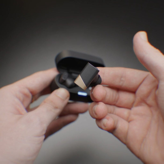 Two hands are holding a pair of Status Audio Pro X True Wireless - GoldenSound Edition earbuds, with one earbud being removed from its open charging case. The background is blurred, drawing focus to the limited-edition IEMs and hands.