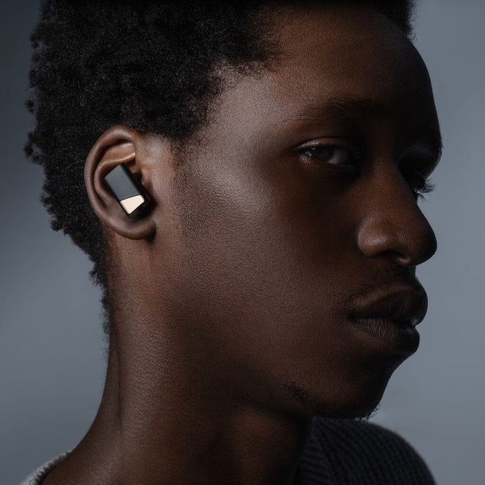 A person with short curly hair wears a Status Audio Pro X True Wireless – GoldenSound Edition earbud in black and gold, looking slightly off to the side against a neutral background.