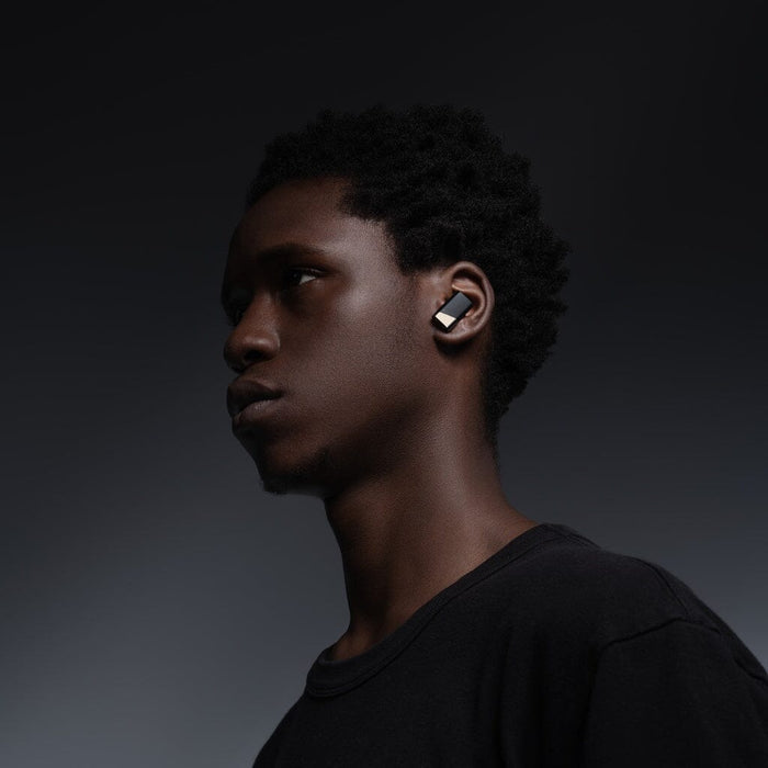 A young person with short curly hair wears a black T-shirt and Status Audio Pro X True Wireless - GoldenSound Edition earbuds, looking to the side against a dark gradient background.
