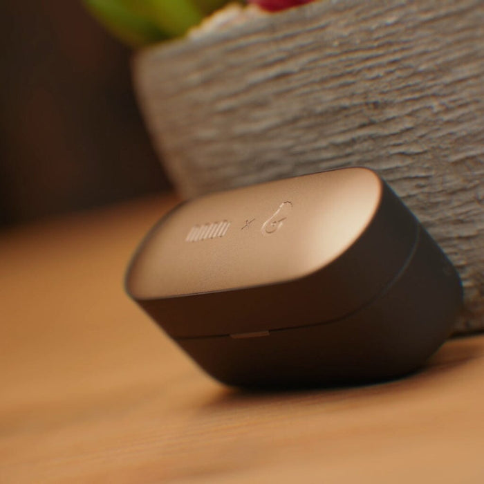 A pair of Status Audio Pro X True Wireless - GoldenSound Edition earbuds in their closed gold and black case rests on a wooden surface, with a textured plant pot softly blurred in the background—an elegant display of this limited-edition release from Status Audio.