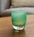 strength teal green hand-blown glass votive candle holder on a wood coffee table with gray couch in background.
