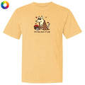 A pale yellow short-sleeve t-shirt with a graphic of a dog wearing sunglasses and a plaid scarf, next to a basket of red apples, with the text 'the dog days of fall' printed below. A small multicolor color wheel icon is in the top-left corner.