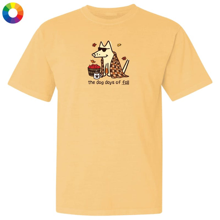 A pale yellow short-sleeve t-shirt with a graphic of a dog wearing sunglasses and a plaid scarf, next to a basket of red apples, with the text 'the dog days of fall' printed below. A small multicolor color wheel icon is in the top-left corner.