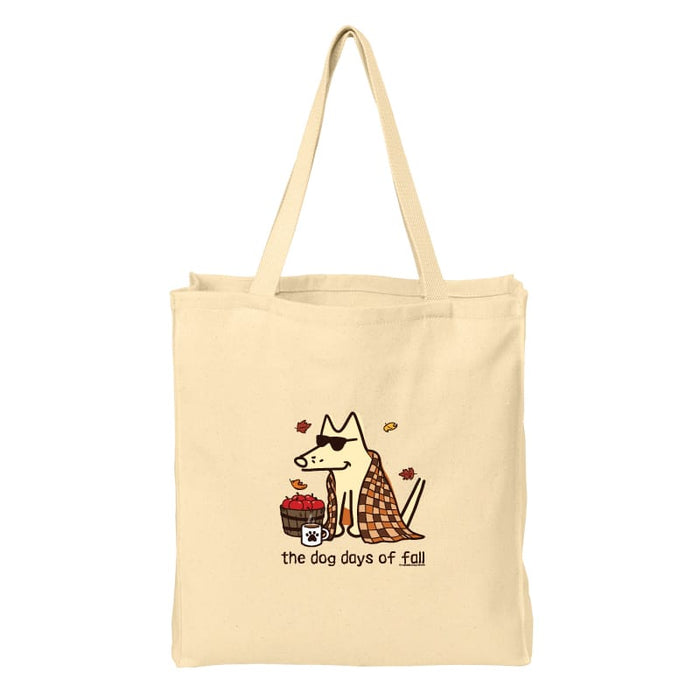 A beige canvas tote bag with a printed graphic of a dog wearing sunglasses and a plaid blanket, next to a basket of red apples, with the text 'the dog days of Fall' below.