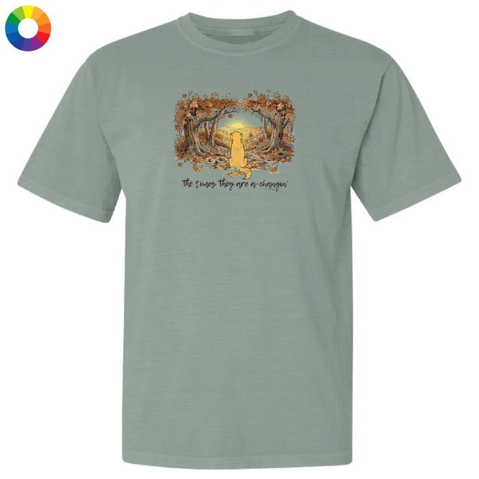 A sage green short-sleeve t-shirt with a graphic of a yellow cat sitting in a forest of trees with autumn leaves, and the text 'The cats, they see or disappear' printed below. A color wheel icon is visible in the top-left corner.